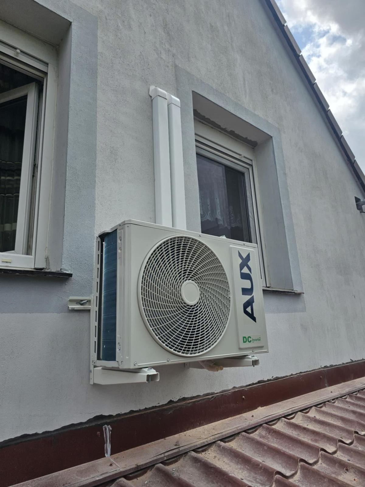 Biała jednostka zewnętrzna klimatyzacji AUX zamontowana na elewacji budynku, widoczna z perspektywy dachu. Rury odprowadzające biegną wzdłuż ściany.