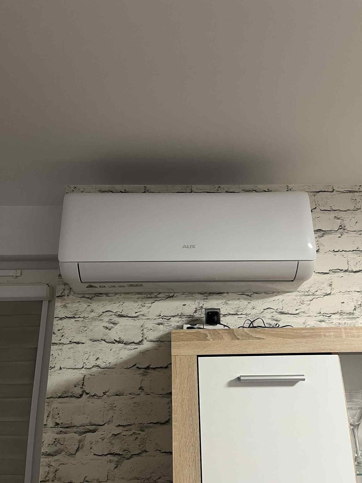 Biały klimatyzator AUX zamontowany na ścianie z cegieł, nad szafką z elementami drewna. Widoczne przewody i czujnik. Oświetlenie górne.