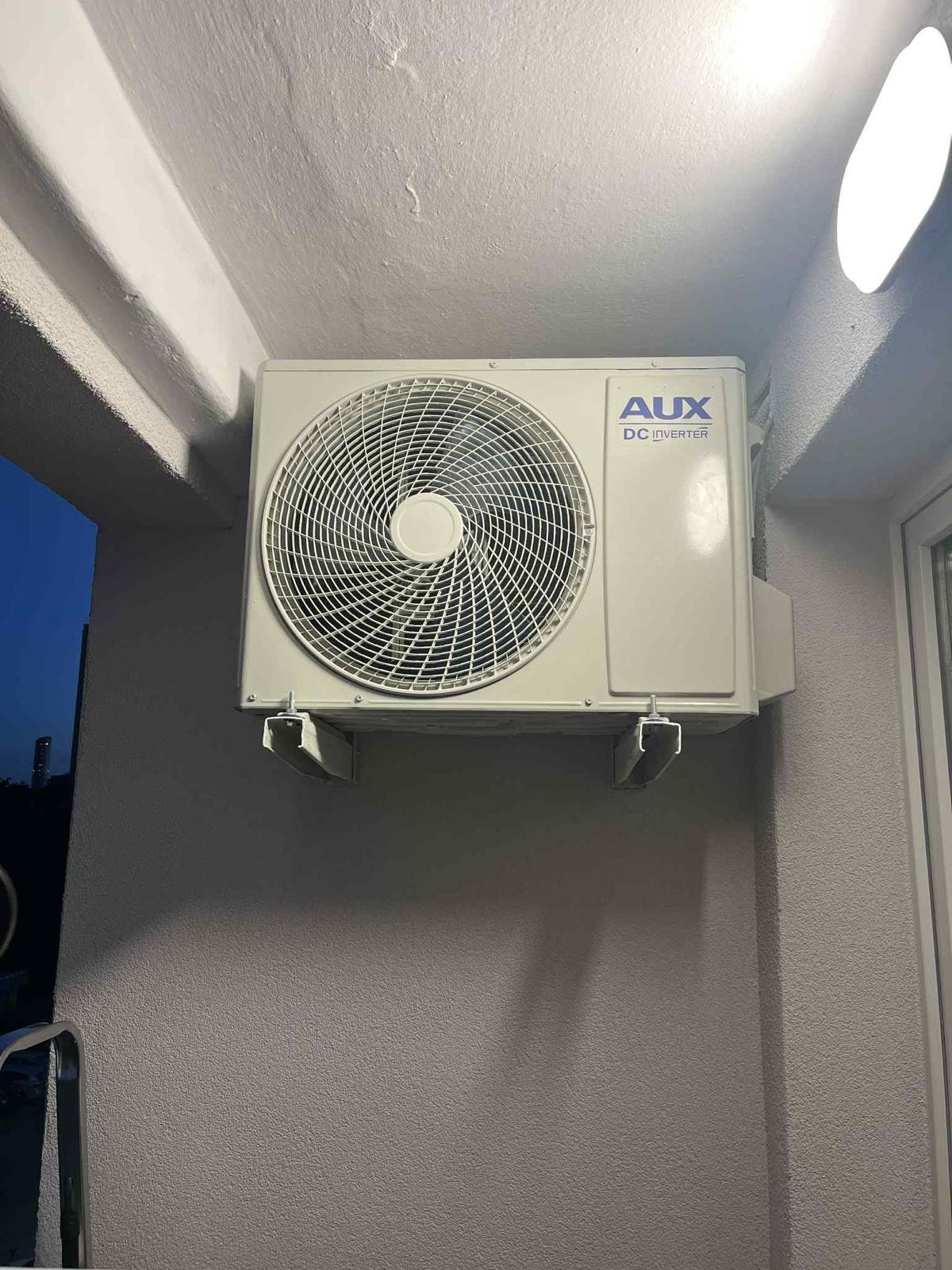 Klimatyzator AUX DC Inverter zamontowany na szarej ścianie zewnętrznej budynku, widoczny z boku, z fragmentem okna po prawej stronie i ciemnym tłem po lewej.
