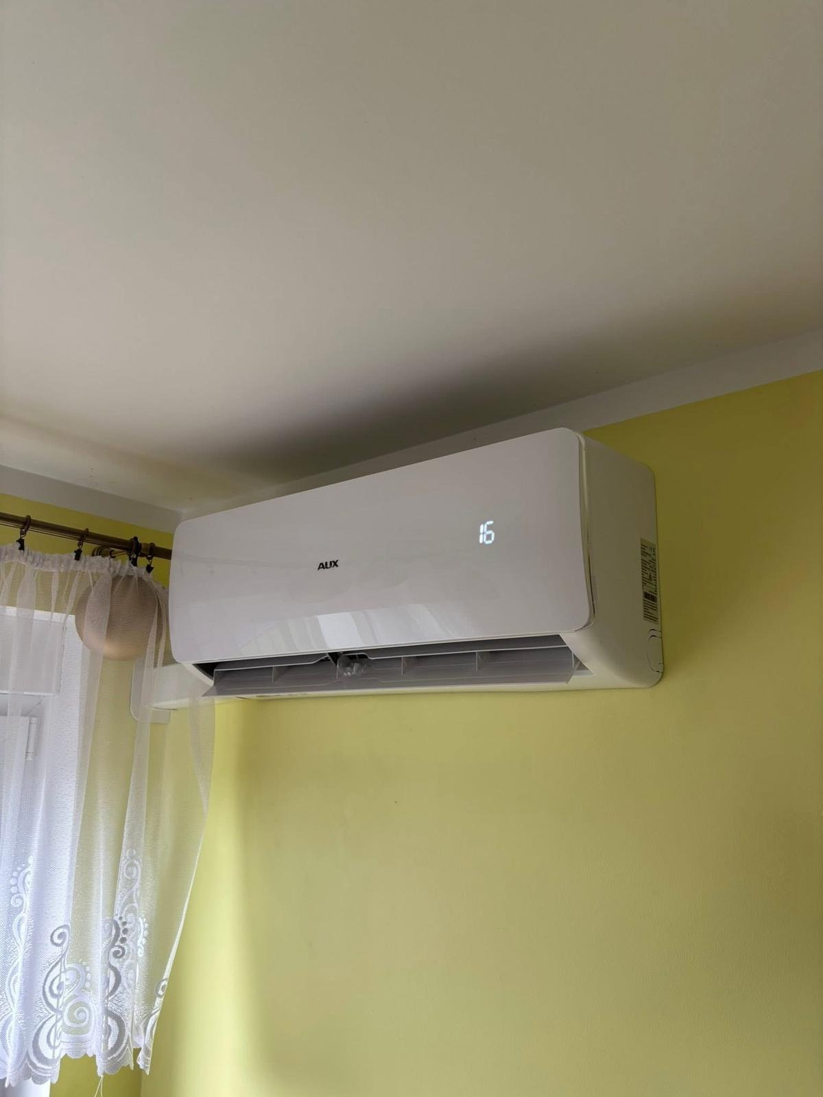 Biały klimatyzator AUX z wyświetlaczem temperatury (16°C) zamontowany na żółtej ścianie, obok okna z firanką. Widok z dołu.