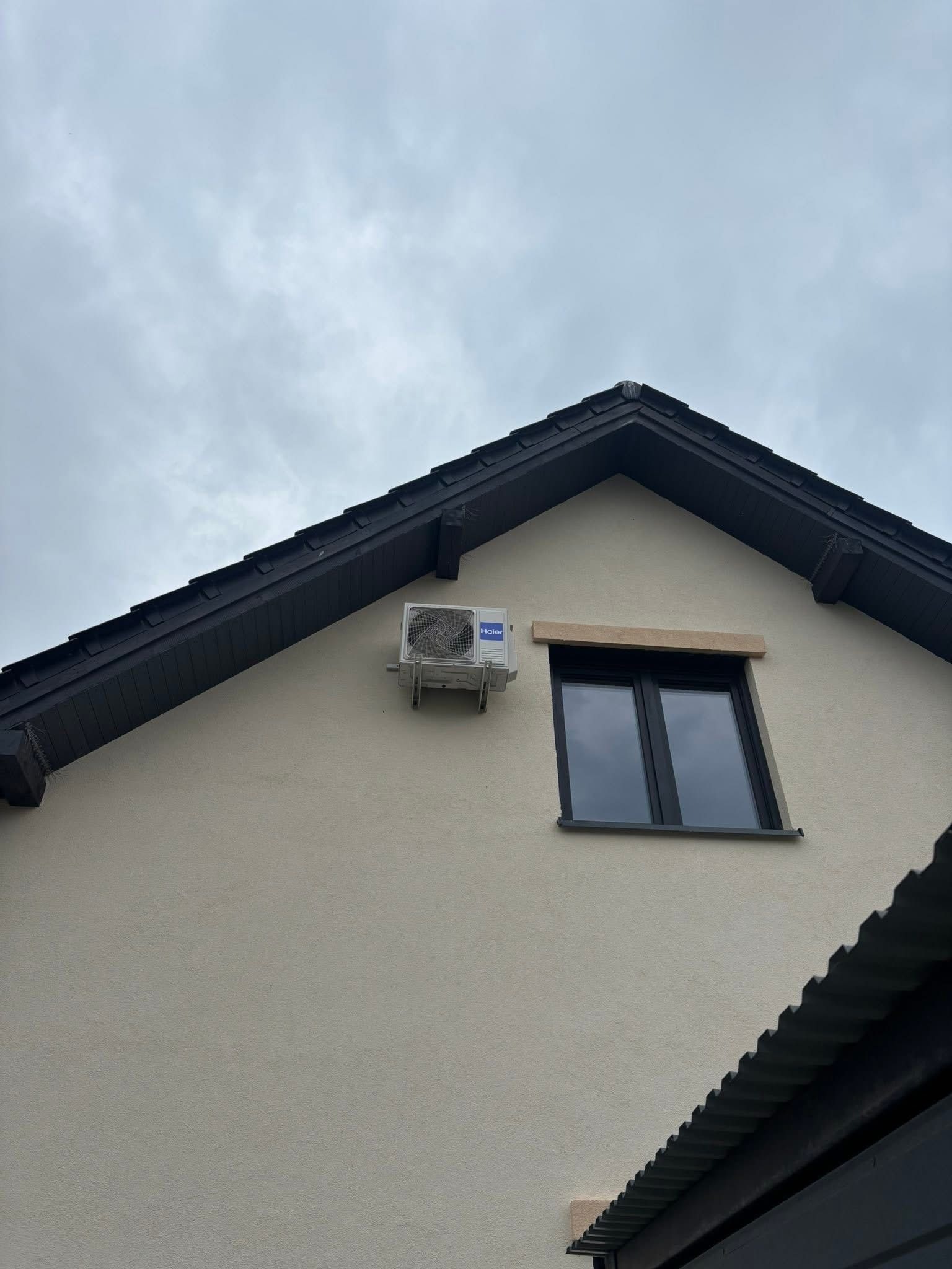Klimatyzator zamontowany na elewacji domu z ciemną dachówką i oknem. Widok z dołu na szarym tle nieba.