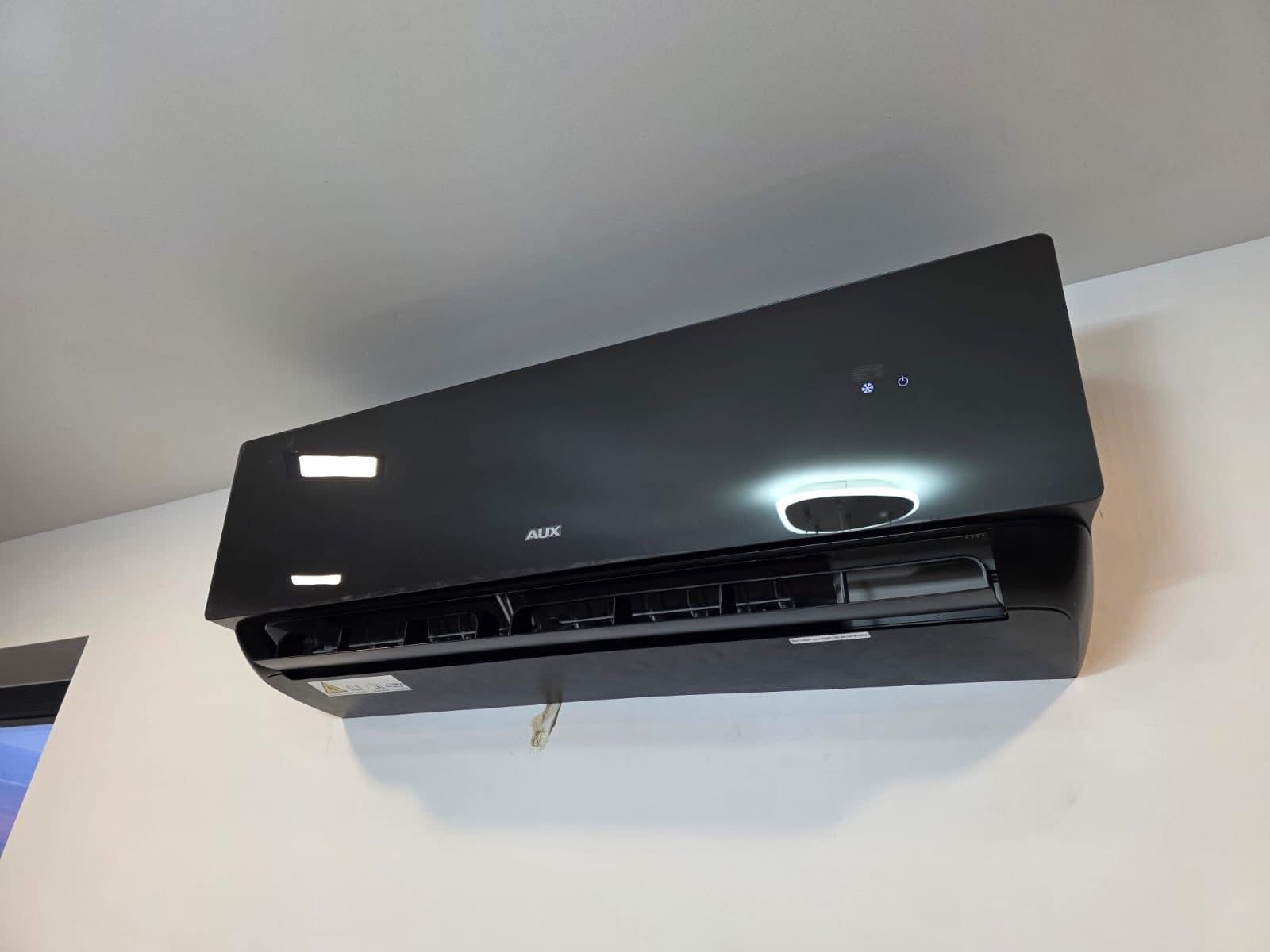 Czarny klimatyzator AUX zamontowany na białej ścianie, widoczne diody sygnalizacyjne i logo producenta. Minimalistyczny design, nowoczesne wnętrze.
