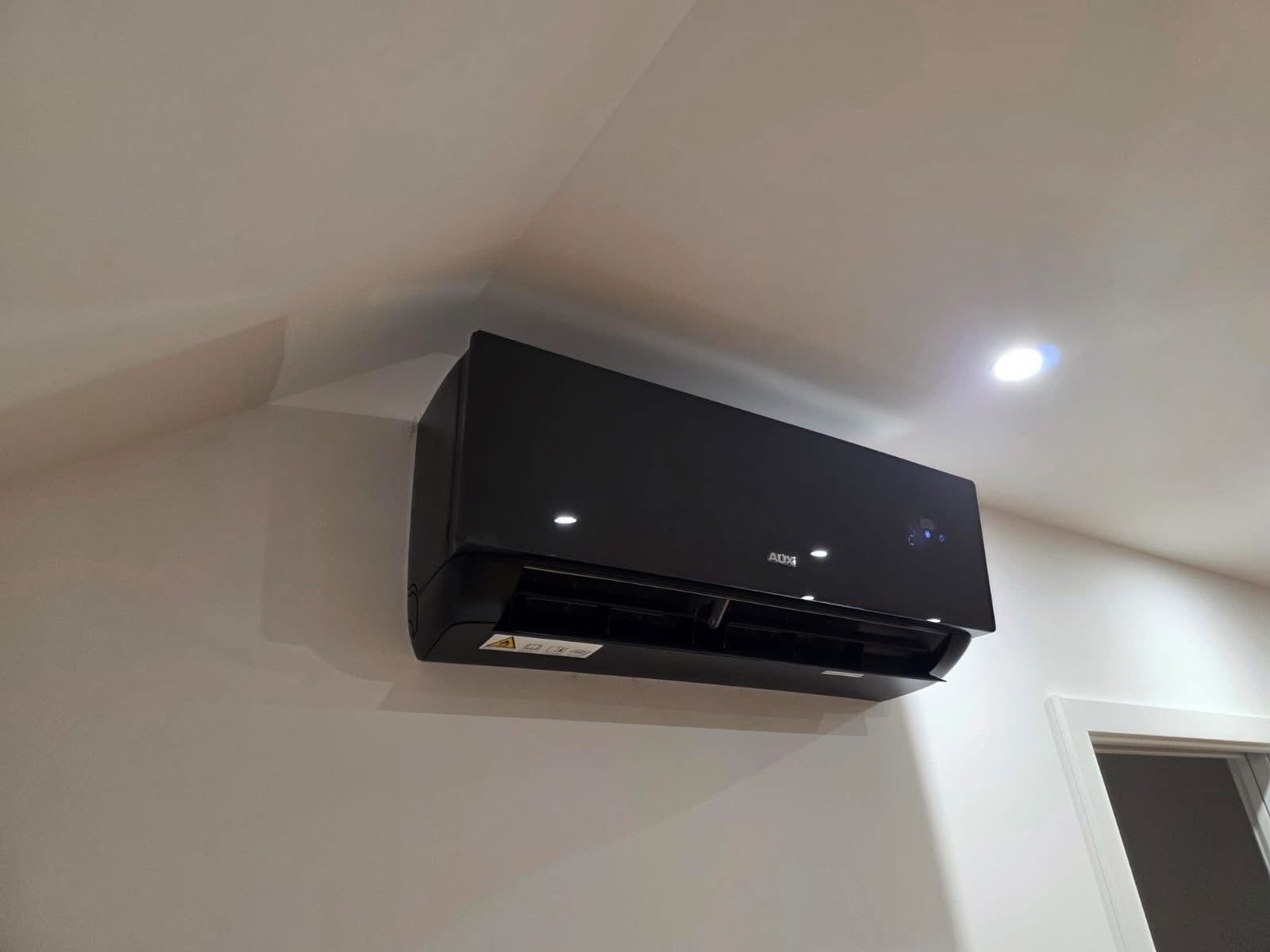 Czarna klimatyzacja AUX, zamontowana na jasnej ścianie w rogu pokoju, widoczna z dołu pod kątem. Minimalistyczny design, nowoczesne wnętrze.