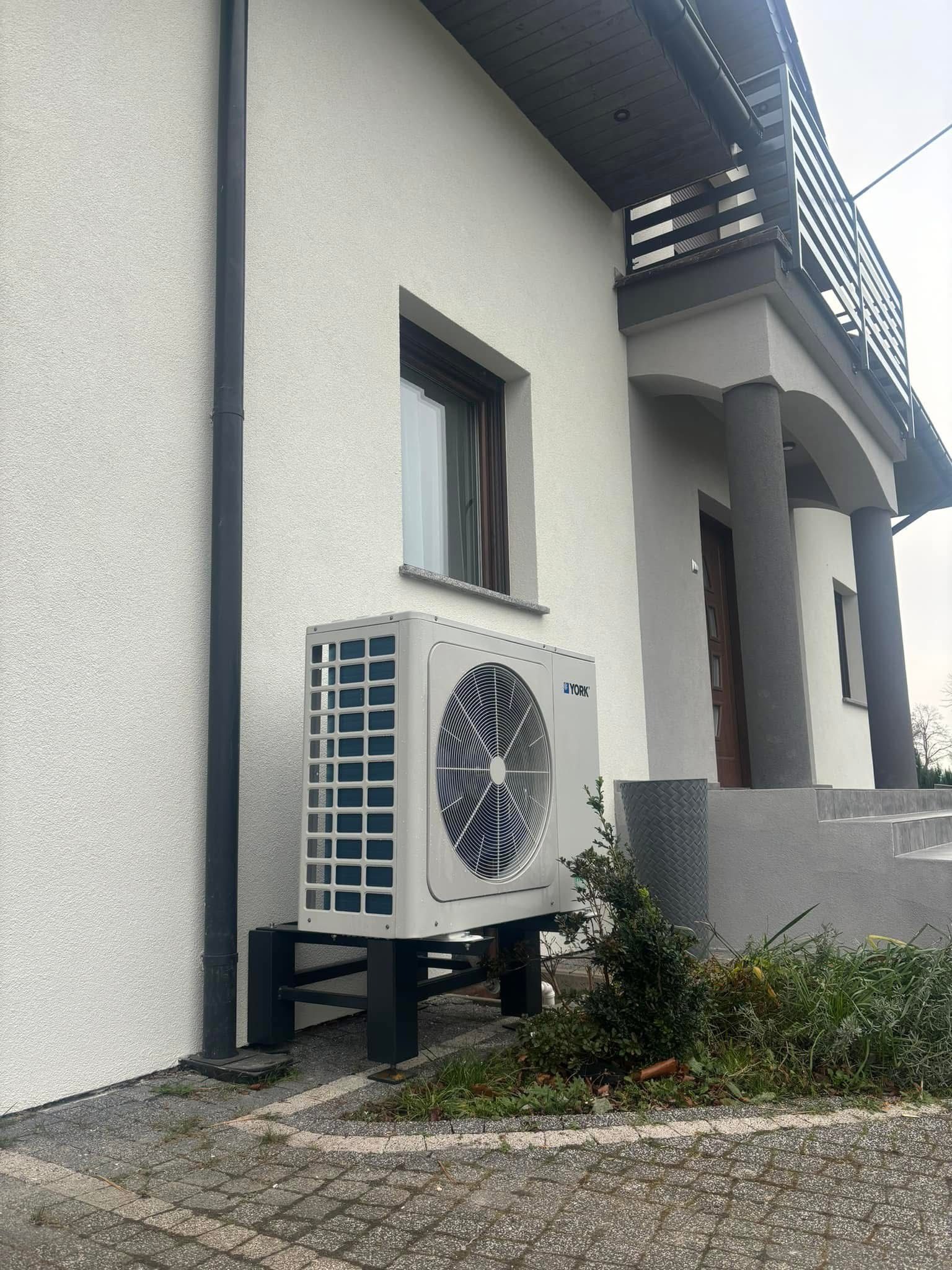 Biała pompa ciepła York na tle jasnej elewacji domu z balkonem i szarą rynną. Widoczne kostki brukowe i fragment zieleni. Ujęcie z dołu.