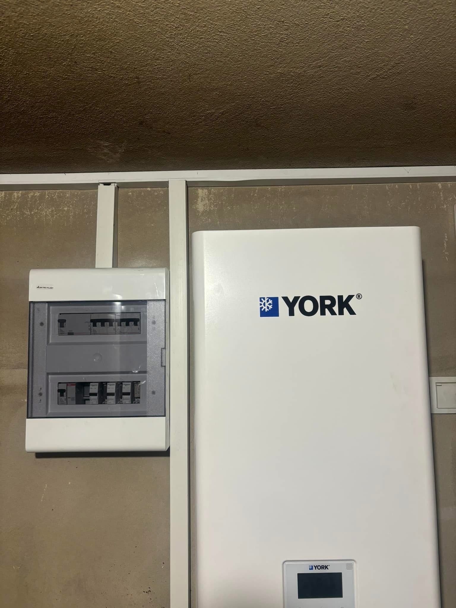 Biała pompa ciepła marki York zamontowana obok skrzynki elektrycznej na ścianie o nierównej fakturze. Widoczne elementy instalacji.