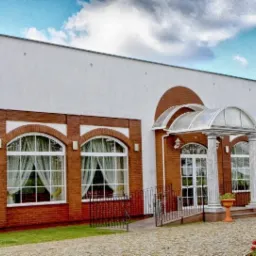 Elewacja budynku z czerwonej cegły z białymi oknami łukowymi i kolumnami przy wejściu, dekoracyjne kwiaty w donicach i kostka brukowa na podjeździe.