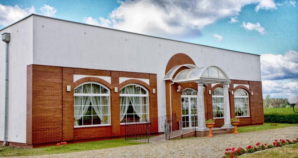 Elewacja budynku z czerwonej cegły z białymi oknami łukowymi i kolumnami przy wejściu, dekoracyjne kwiaty w donicach i kostka brukowa na podjeździe.