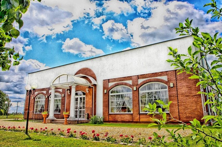 Elewacja budynku z cegły z białym tynkiem, kolumnami przy wejściu, dużymi oknami z białymi firankami i zadbanym ogrodem z czerwonymi kwiatami.