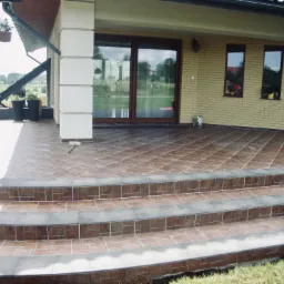 Wyłożony brązowymi płytkami taras z trzema stopniami, widok na jasną elewację domu z oknami i kwiatami, oraz fragment krajobrazu w tle.