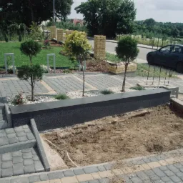 Ułożona kostka brukowa na schodach i podwyższonym klombie z nasadzeniami, widoczny kabel elektryczny w ziemi, w tle trawnik i zaparkowany samochód.