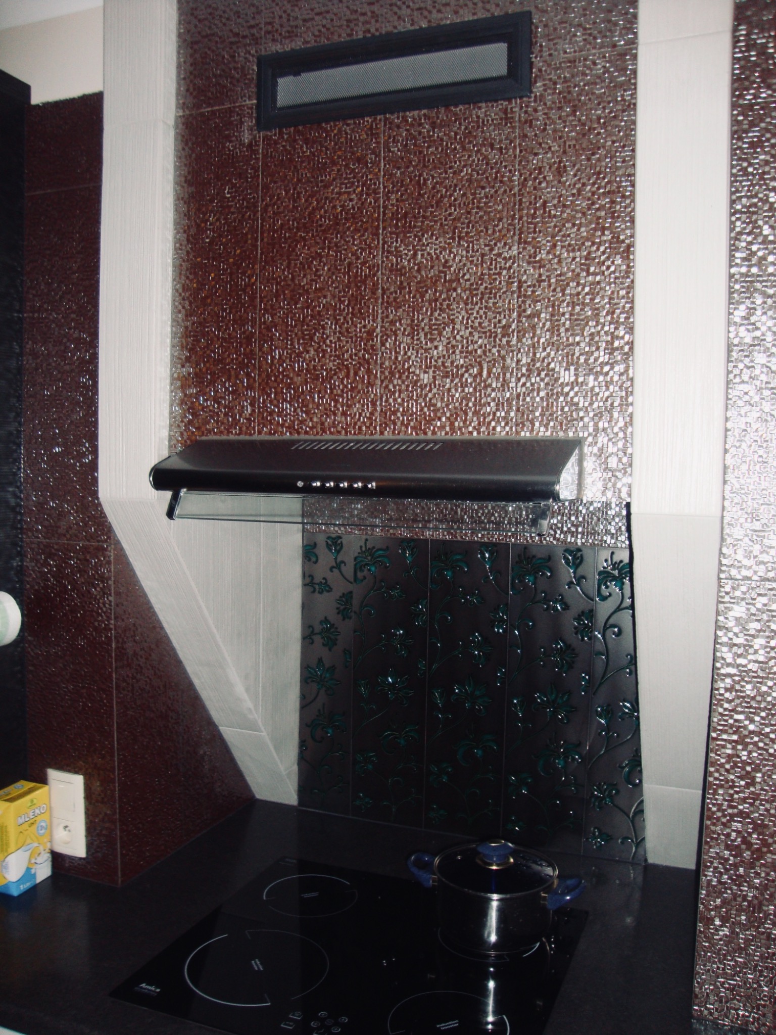 Kuchenna ściana nad płytą indukcyjną wykończona błyszczącymi brązowymi i czarnymi płytkami z motywem kwiatowym, pod okapem; na płycie stoi garnek z niebieskimi uchwytami.