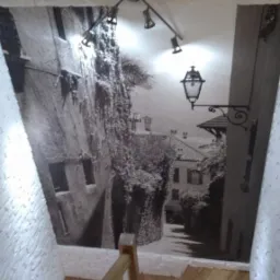 Fototapeta z motywem włoskiej uliczki na ścianie klatki schodowej, oświetlona lampą sufitową z trzema reflektorami, widoczne drewniane schody i imitacja cegły na ścianie.