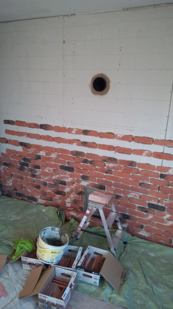 Montaż dekoracyjnych paneli imitujących cegłę na ścianie z otworem, widoczna drabina, wiadro z klejem i kartony z panelami.