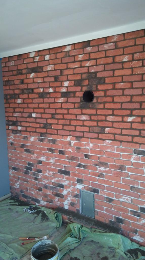 Ściana z imitacji cegły z otworem wentylacyjnym i metalową klapką rewizyjną, częściowo zakryta folią ochronną i zabrudzona farbą, z widocznymi narzędziami malarskimi.