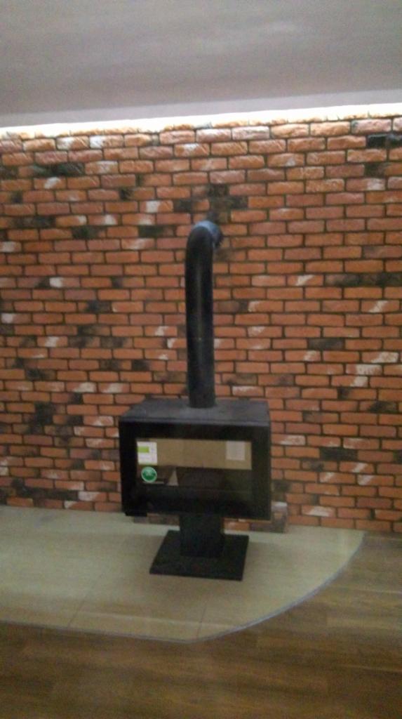 Ciemny piec wolnostojący z czarną rurą dymową na tle ściany wyłożonej dekoracyjną cegłą imitującą starą czerwoną cegłę z ciemnymi przebarwieniami, podświetlonej listwą LED.