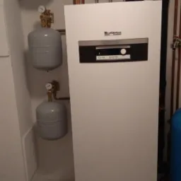 Biała jednostka grzewcza Buderus z podłączonymi miedzianymi rurami, zbiornikami wyrównawczymi i manometrami w kotłowni z ceramiczną podłogą.