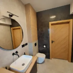 Nowoczesna łazienka z owalnym lustrem, białą umywalką na drewnianym blacie, toaletą i drewnianymi drzwiami z wzorem jodełki, wykończona szarymi płytkami.