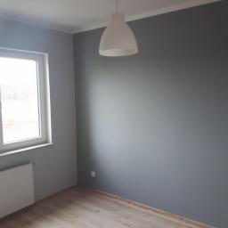 Group Service PL - Pomalowane na szaro wnętrze pokoju z drewnianą podłogą, białym grzejnikiem i lampą wiszącą w kształcie stożka, widok na okno z zewnątrz.