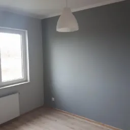 Pomalowane na szaro wnętrze pokoju z drewnianą podłogą, białym grzejnikiem i lampą wiszącą w kształcie stożka, widok na okno z zewnątrz.