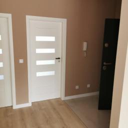 Group Service PL - Korytarz po remoncie: ściany w odcieniach beżu, białe drzwi z poziomymi przeszkleniami, ciemne drzwi wejściowe, podłoga z paneli imitujących drewno, listwy przypodłogowe.
