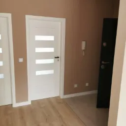 Korytarz po remoncie: ściany w odcieniach beżu, białe drzwi z poziomymi przeszkleniami, ciemne drzwi wejściowe, podłoga z paneli imitujących drewno, listwy przypodłogowe.