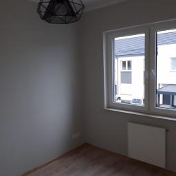 Group Service PL - Pomalowane na szaro wnętrze pokoju z drewnianą podłogą, białym grzejnikiem pod oknem i wiszącą czarną, geometryczną lampą.