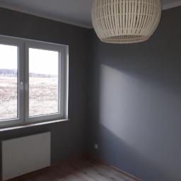 Group Service PL - Szare wnętrze pokoju po odświeżeniu: pomalowane ściany, drewnopodobna podłoga, biały grzejnik pod oknem, pleciona lampa w stylu boho.