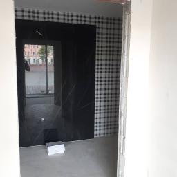 Group Service PL - Wnętrze łazienki w trakcie prac wykończeniowych: czarna płyta imitująca marmur na ścianie, wzorzysta tapeta w kratę, surowy ościeżnica i widok na budynki przez okno.