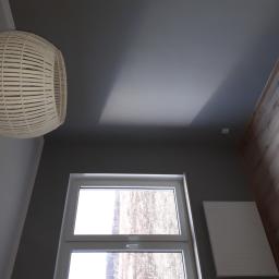 Group Service PL - Wnętrze pokoju po odświeżeniu: szara ściana, białe okno z widokiem na zewnątrz, drewniana podłoga i pleciona lampa wisząca w rogu kadru.