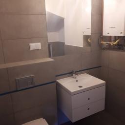 Group Service PL - Nowoczesna łazienka z umywalką nablatową na szafce z dwiema szufladami, lustrem i grzejnikiem gazowym Viessmann na ścianie wyłożonej płytkami w odcieniach szarości z subtelnym niebieskim akcentem.