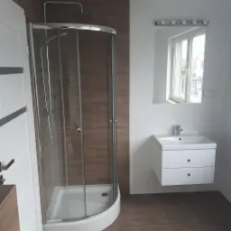 Nowoczesna łazienka po remoncie z kabiną prysznicową z chromowanymi elementami, białą szafką pod umywalkę z dwoma szufladami i dużym lustrem z oświetleniem punktowym.