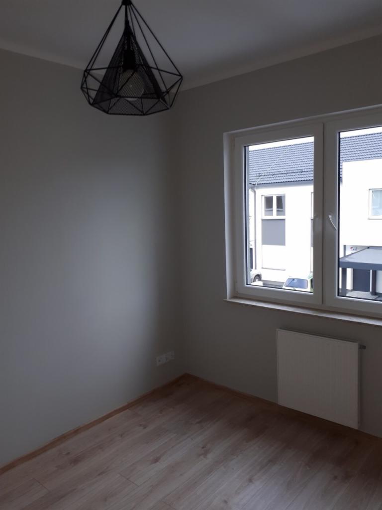 Pomalowane na szaro wnętrze pokoju z drewnianą podłogą, białym grzejnikiem pod oknem i wiszącą czarną, geometryczną lampą.