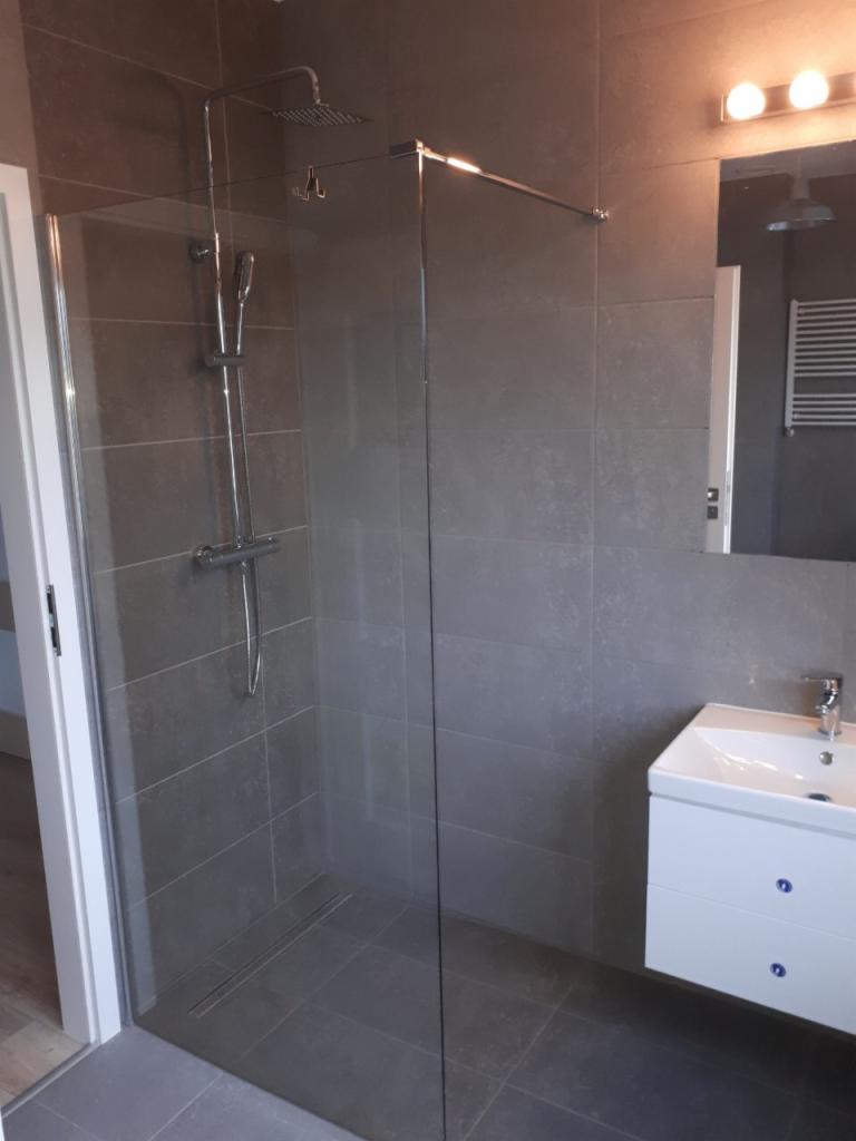 Nowoczesna kabina prysznicowa typu walk-in z szarymi płytkami, chromowaną armaturą i odpływem liniowym w podłodze, uzupełniona białą szafką z umywalką i oświetleniem nad lustrem.
