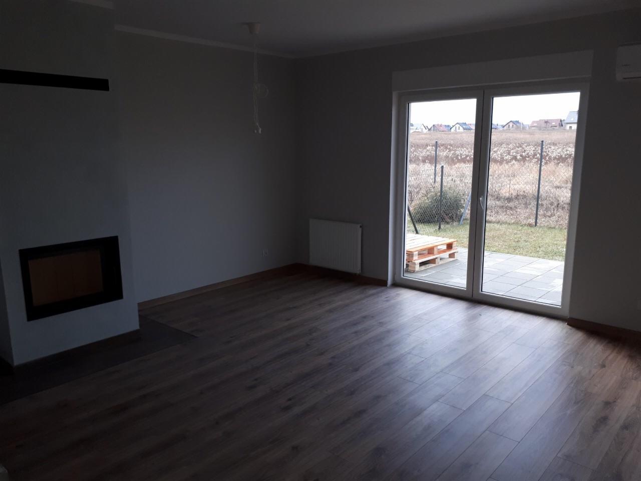 Wnętrze pokoju z panelami podłogowymi w odcieniach brązu, kominkiem z czarną obudową i dużymi przesuwnymi drzwiami balkonowymi wychodzącymi na ogród z widoczną paletą na tarasie.