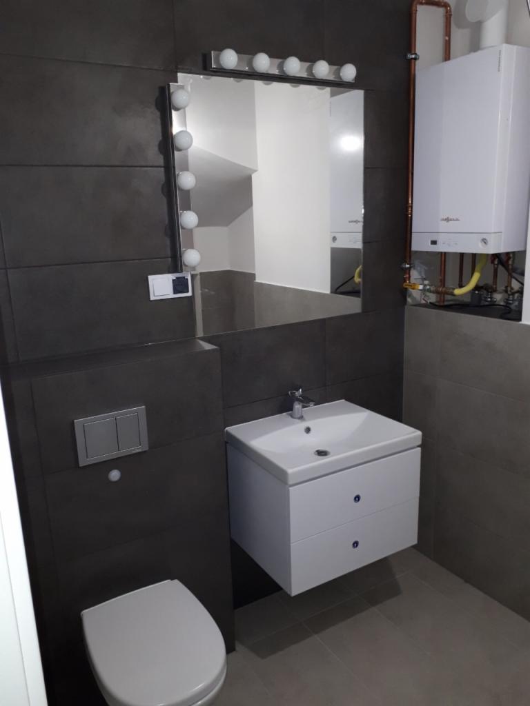Nowoczesna łazienka z szarą płytką, wiszącą szafką z umywalką, toaletą i dużym lustrem z oświetleniem punktowym oraz piecem grzewczym.