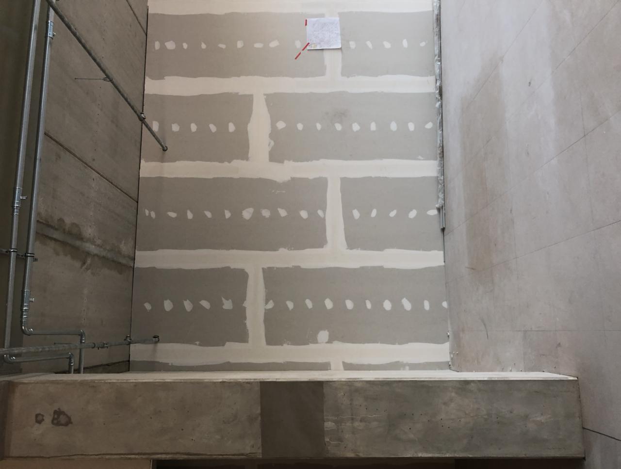 Ściana w trakcie montażu płyt gipsowo-kartonowych, widoczne łączenia i miejsca po wkrętach, zarys instalacji rurowej po lewej stronie i fragment ściany wyłożonej płytkami po prawej.