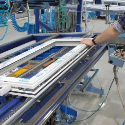 Proces produkcji ramy okiennej na linii montażowej z elementami pneumatycznymi i elektrycznymi, widoczna ręka pracownika w kombinezonie.