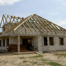 Wznoszenie domu z białego pustaka: widoczne ściany, otwory okienne i drewniana konstrukcja dachu w trakcie montażu, na tle błękitnego nieba.