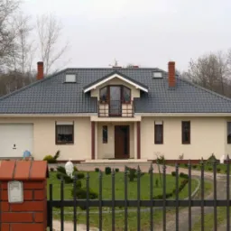 Dom jednorodzinny z szarym dachem z dachówki ceramicznej, widoczne okna dachowe i kominy. Elewacja w kolorze beżowym, brama garażowa biała.