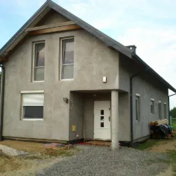 Surowy, otynkowany dom jednorodzinny z szarą elewacją, widoczne okna z roletami, kolumna podtrzymująca zadaszenie nad wejściem, teren wokół domu w trakcie prac wykończeniowych.