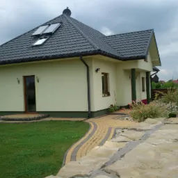 Gotowy dom jednorodzinny z szarą dachówką, panelami słonecznymi i jasną elewacją, otoczony trawnikiem i ścieżką z kostki brukowej, z widocznym fragmentem kamiennego murku ogrodzeniowego.