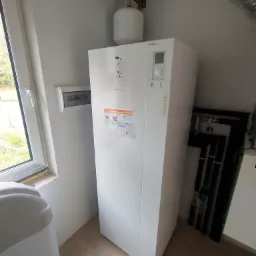 Biała pompa ciepła Atlantic z widocznym manometrem na górze, zamontowana w pomieszczeniu z beżową podłogą, obok okna i przewodów instalacyjnych.