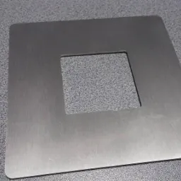 Kwadratowa, metalowa płyta z wyciętym kwadratowym otworem pośrodku, leżąca na szarej, teksturowanej powierzchni.