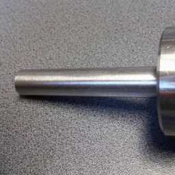 Walcowany element metalowy z cylindrycznym trzpieniem i kołnierzem, leżący na szarej powierzchni o drobnej teksturze.