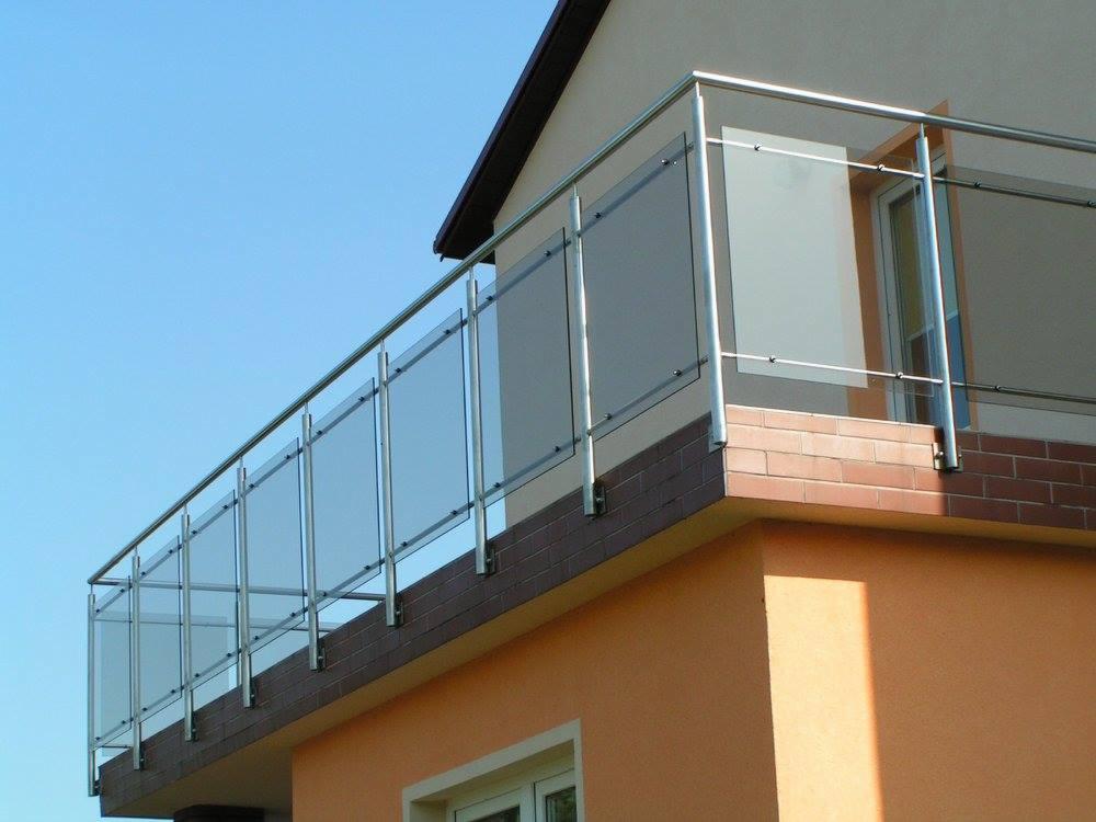 Nowoczesna balustrada balkonowa ze stali nierdzewnej i szkła na tle błękitnego nieba, zamontowana na ceglanym murku pomalowanym na pomarańczowo.