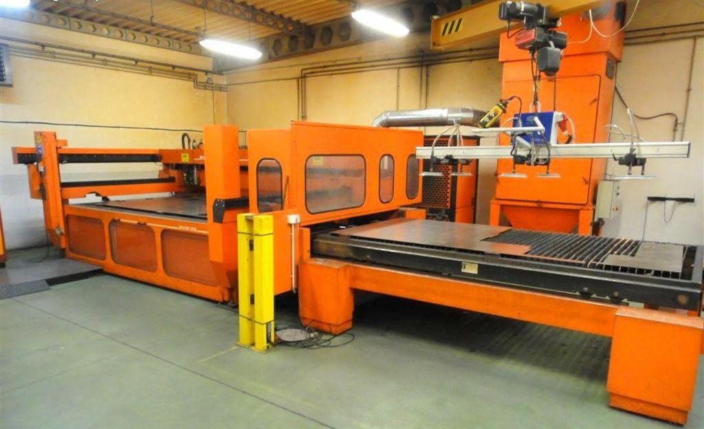 Pomarańczowa maszyna do precyzyjnego cięcia blach z widocznymi elementami mocującymi i systemem odciągowym, na podłodze leżą wycięte elementy.