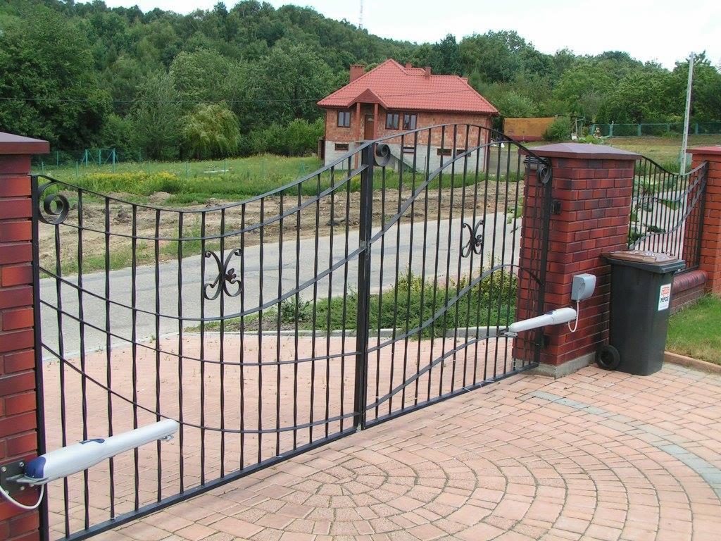 Kuta brama wjazdowa z automatycznym napędem, zamontowana na ceglanych słupkach, prowadząca do posesji z domem o czerwonym dachu.