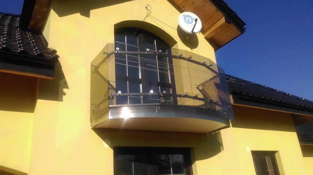 Żółta elewacja budynku z zaokrąglonym balkonem z balustradą ze szkła i metalu, mocowaną punktowo, widok z dołu w słoneczny dzień.