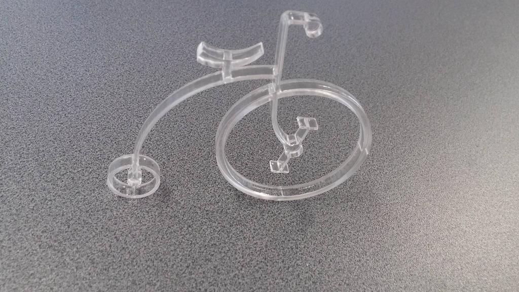 Przezroczysty model roweru typu bicykl wykonany z tworzywa sztucznego, wycięty prawdopodobnie metodą CNC, leżący na szarym, teksturowanym tle.