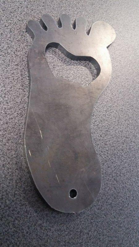 Metalowy otwieracz do butelek w kształcie stopy z wyciętym sercem, wykonany w Zgłobicach, leżący na szorstkiej powierzchni.